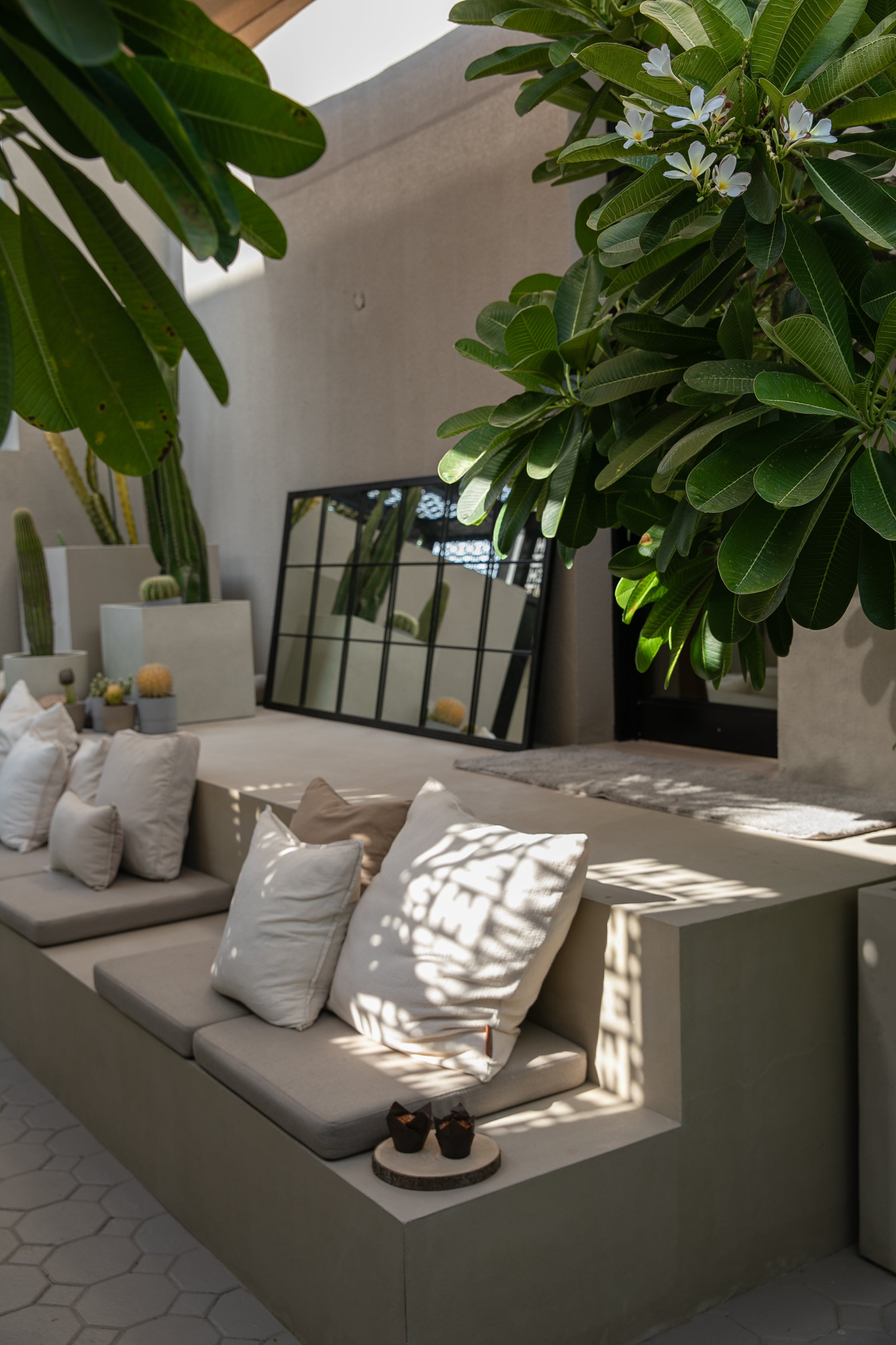 Modern courtyard outdoor step seating with a linen and black decor, decorative fluffy pillows, and a mirror on the linen wall and large frangipani plants with flowers.
