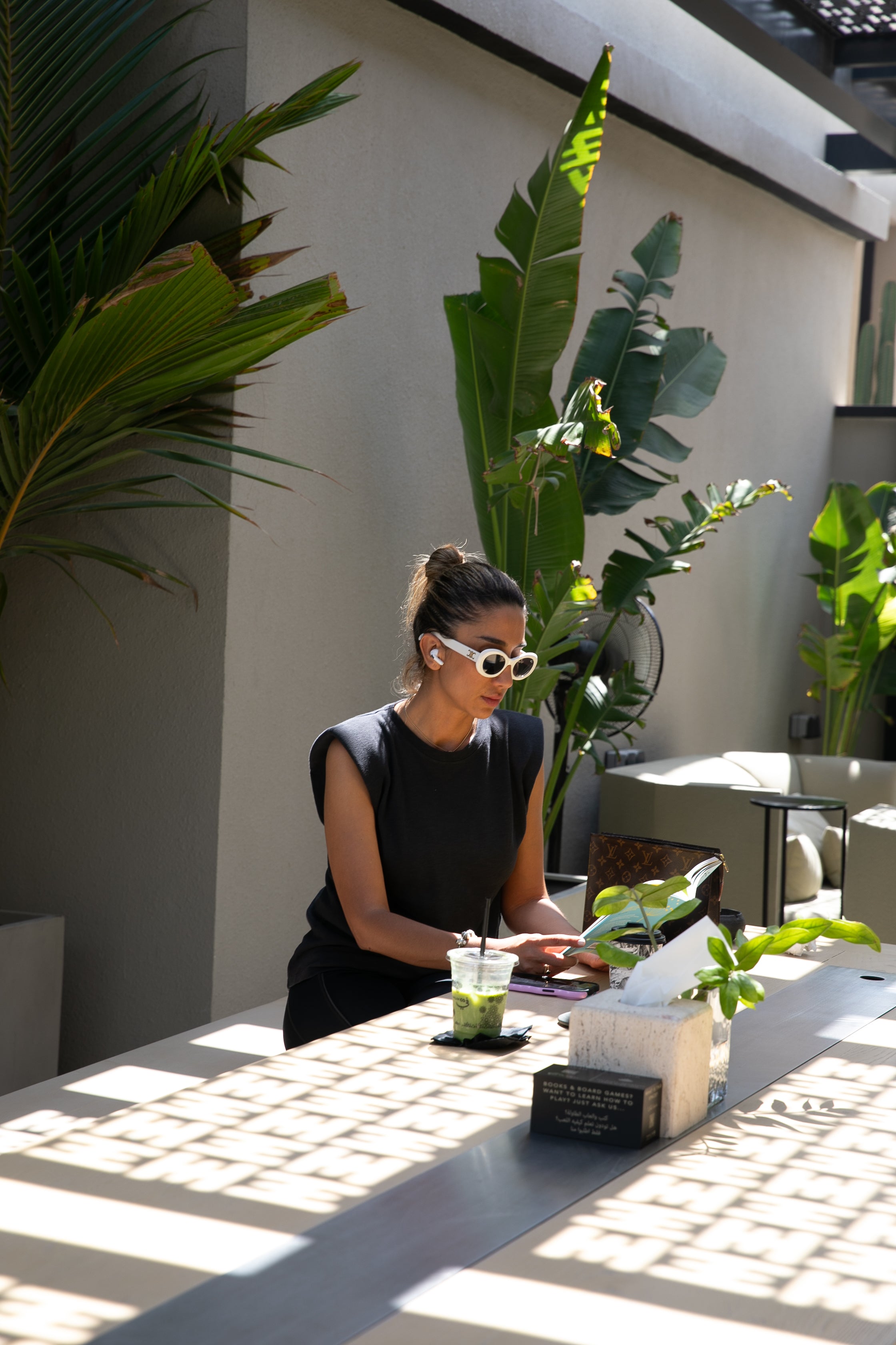 Woman sitting at a table working at Halm Collective Wellness Club enjoying a matcha surrounded by lush greenery and travellers palm with a breezy outdoor fan.