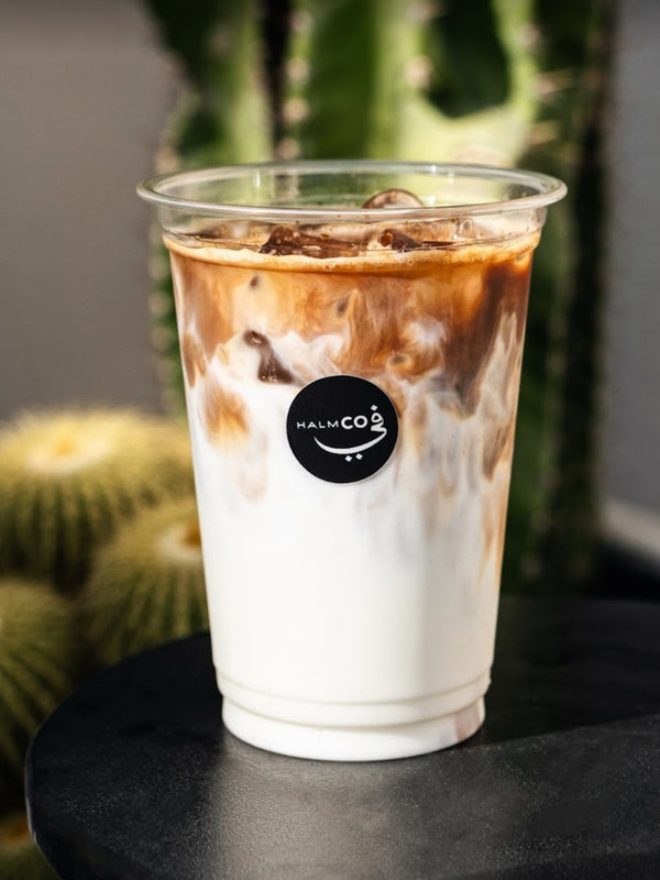 Iced coffee in a clear plastic cup with a logo on a dark surface, cactus in the background.