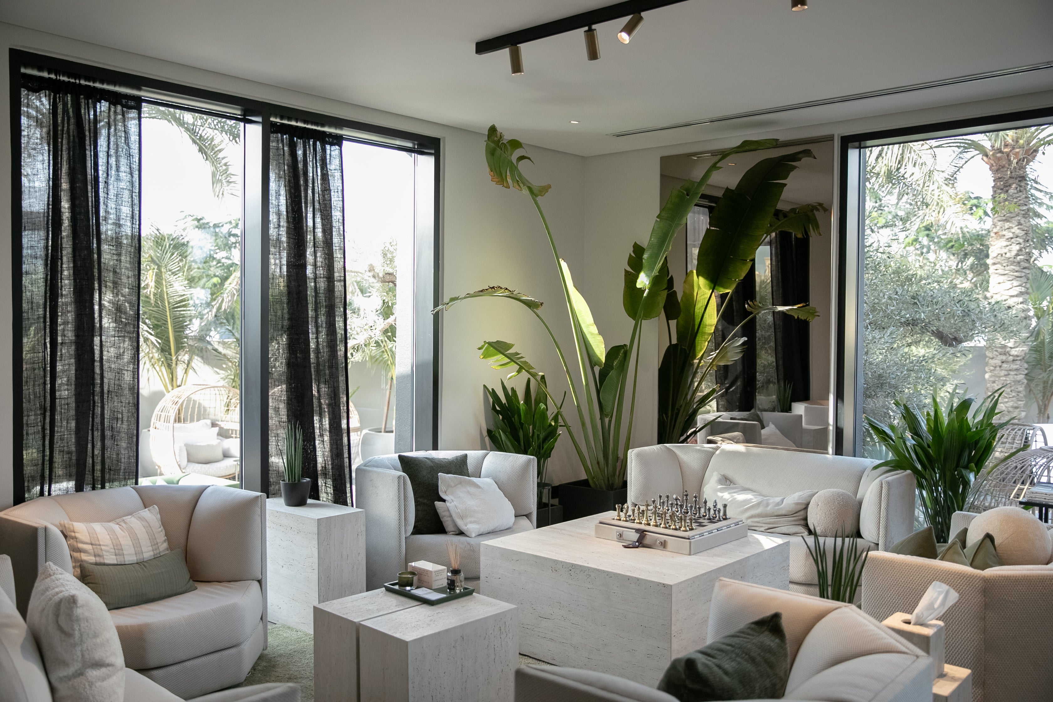 Modern lounge with large windows, indoor tropical plants, neutral-toned armchairs, and a central table with a Halm bespoke leather cream and black chess set, creating a serene and inviting atmosphere