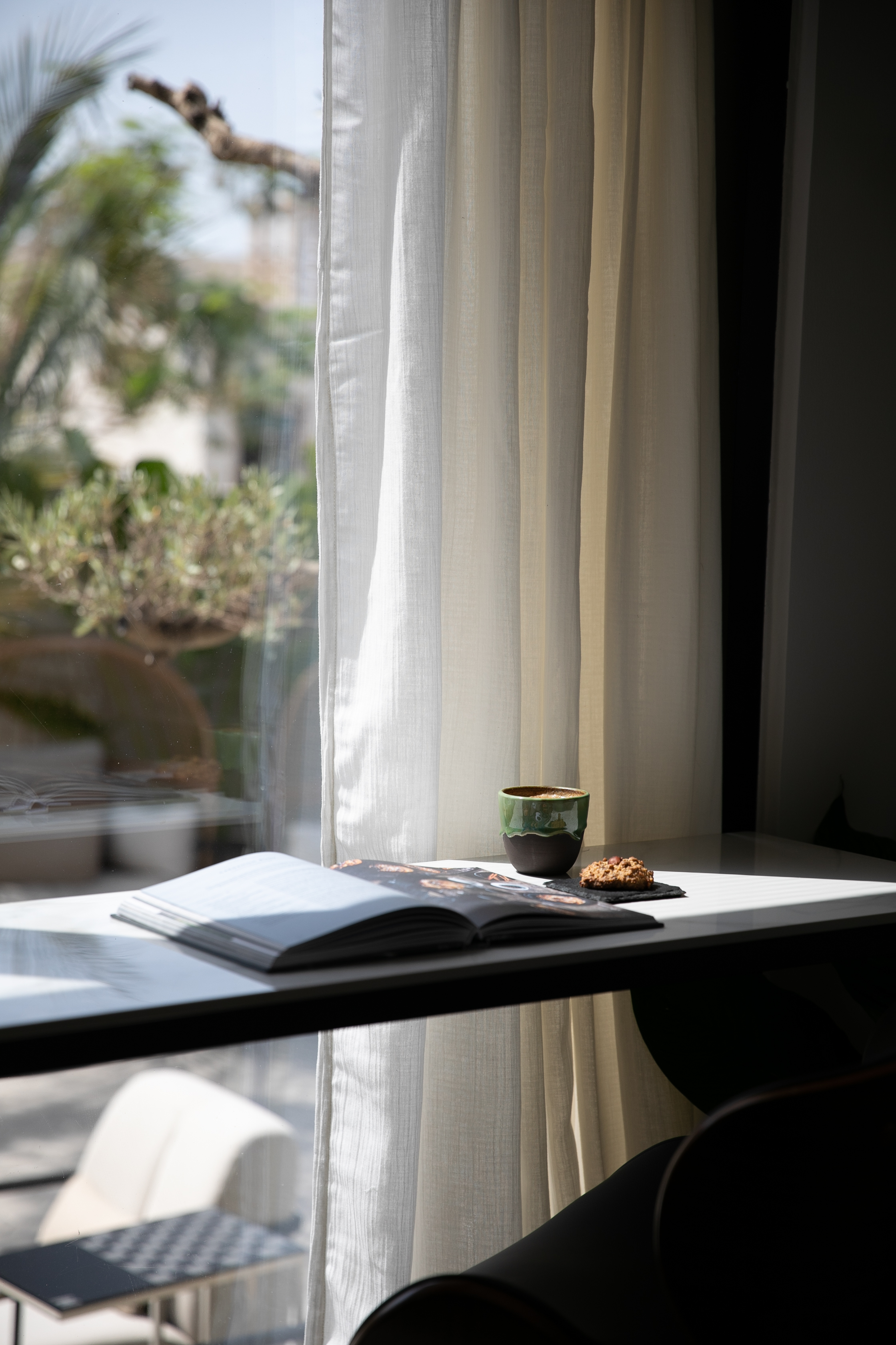 Open book on a table by the window with the green exterior with a bespoke ceramic cup, made in the UAE filled with speciality coffee and nutritious cookie from the Halm Cafe, 705 Jumeirah Beach Road.