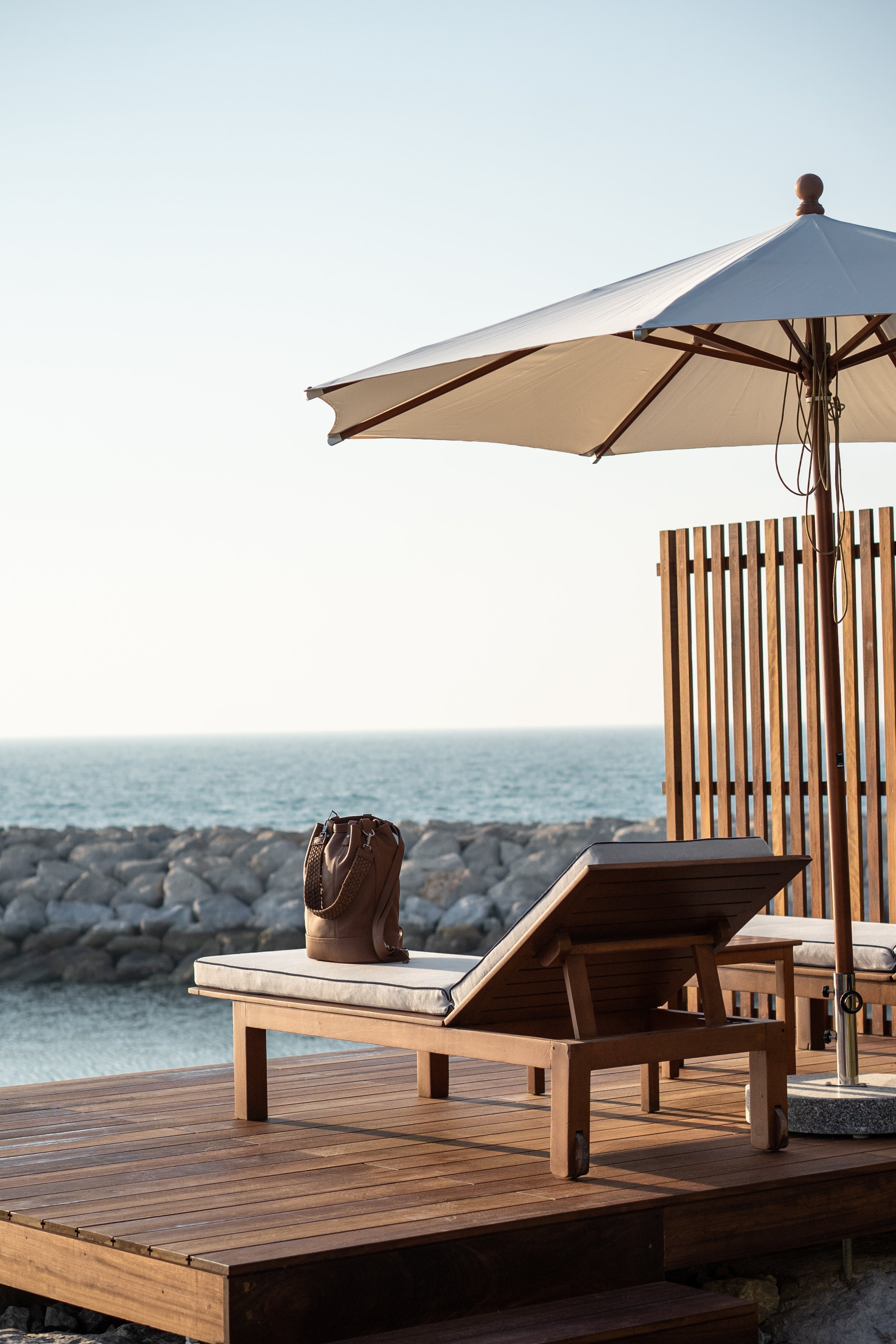 Seaside modern sleek wooden sun lounge with a limited edition Halm leather tote bag, on a jetty with a linen umbrella and ocean view with rocks. 