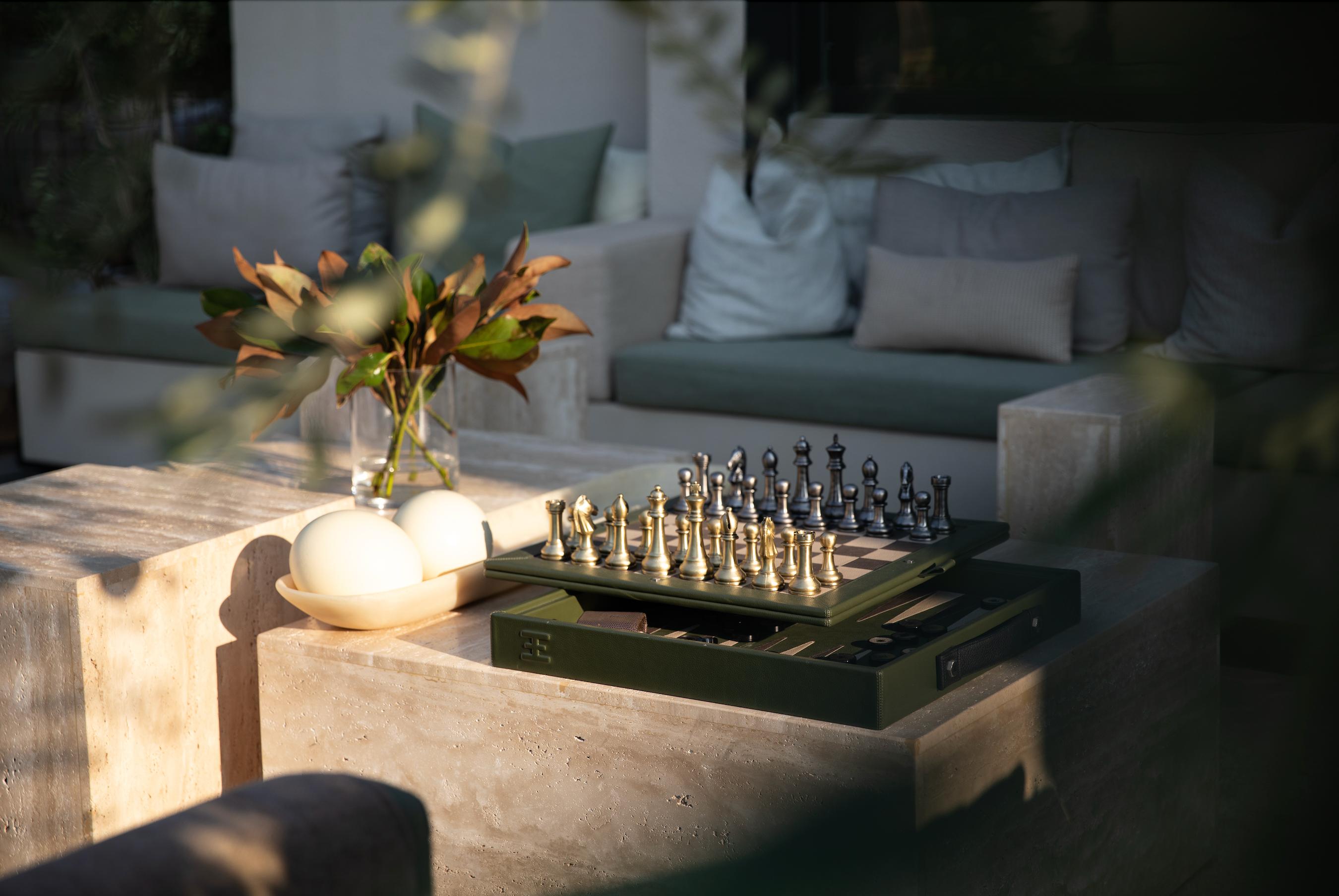 Bespoke Halm green leather chess and backgammon board game on a beige travertine coffee table, a vase of magnolia branches and a designer tray with beige ostrich eggs  with green majlis seating and plush pillows in an olive grove at Halm Collective Wellness Club, Umm Suqeim. 