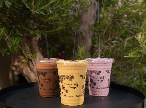 A selection of protein shakes from the Halm Cafe menu in Halm takeaway cups agaisnt a green foliage background.