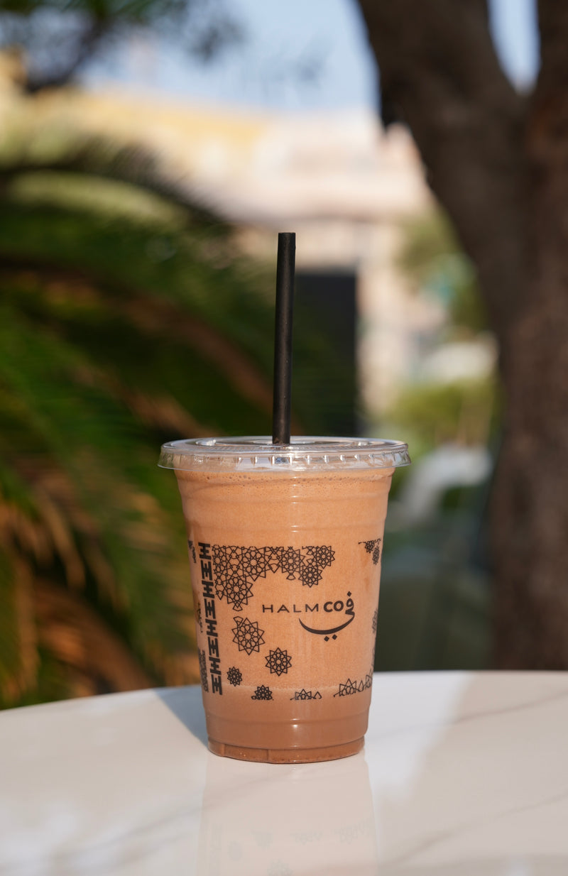 Iced coffee in a clear Halm Coffee cup with a black lid and straw on a white surface, blurred greenery in the background