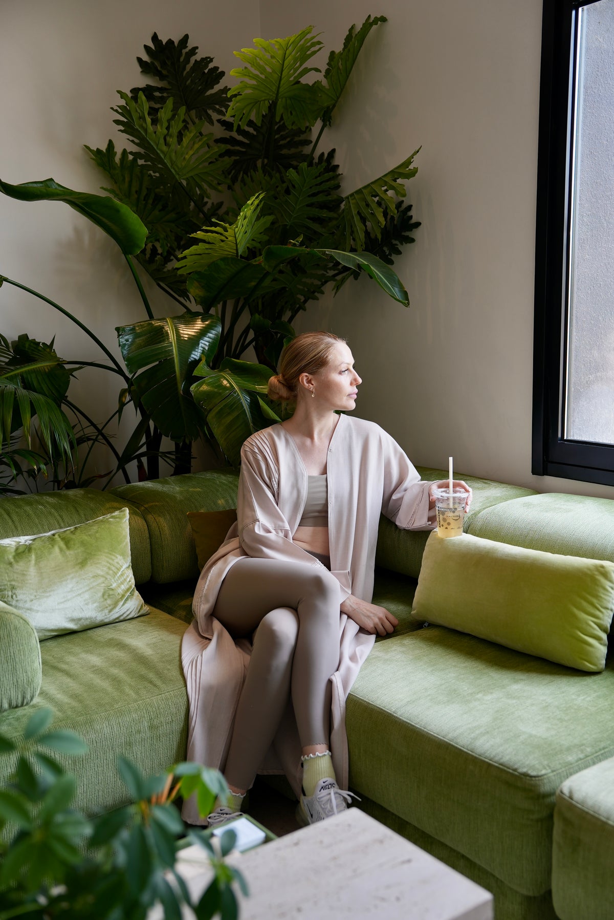 Woman sitting on a green couch with plants around, holding a drink from HALM cafe.