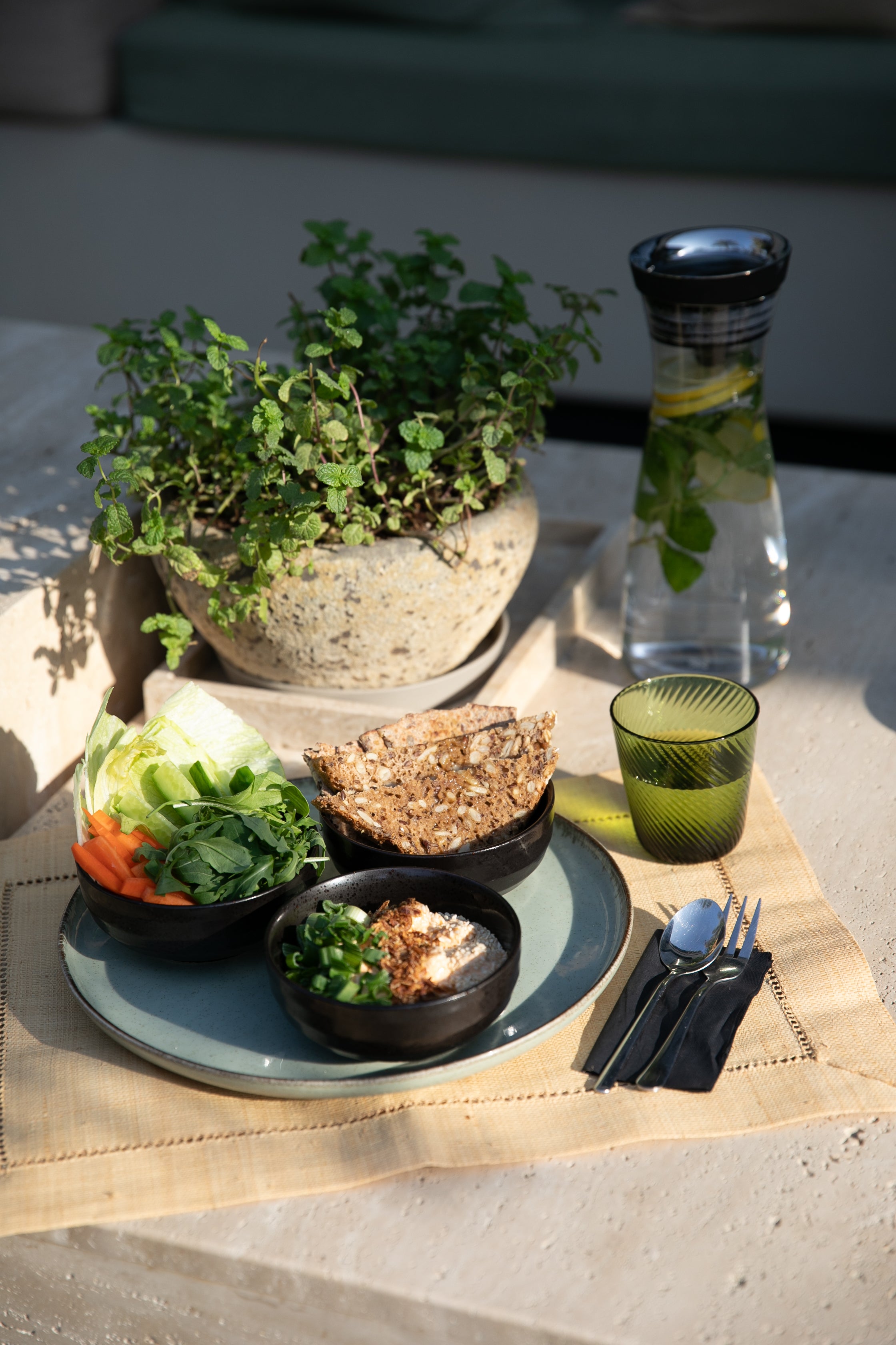Outdoor setting with a healthy high protein meal on a tablecloth and a clay pot of mint herbs including a bowl of salad, bread, and refreshing jug of lemon and herb water.