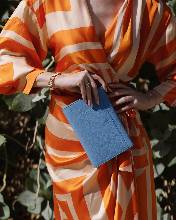 Lady wearing an orange and white striped dress holding a sky blue clutch made in the UAE by Halm limited edition leather bags. 