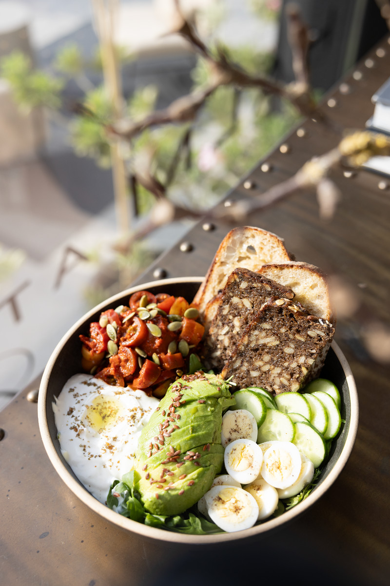 Nutritious colourful  healthy bowl with avocado, boiled eggs, cucumbers, yogurt, wholegrain sourdough bread. Served at an outdoor table with a green plant at Halm Collective Wellness Club in Dubai. 
