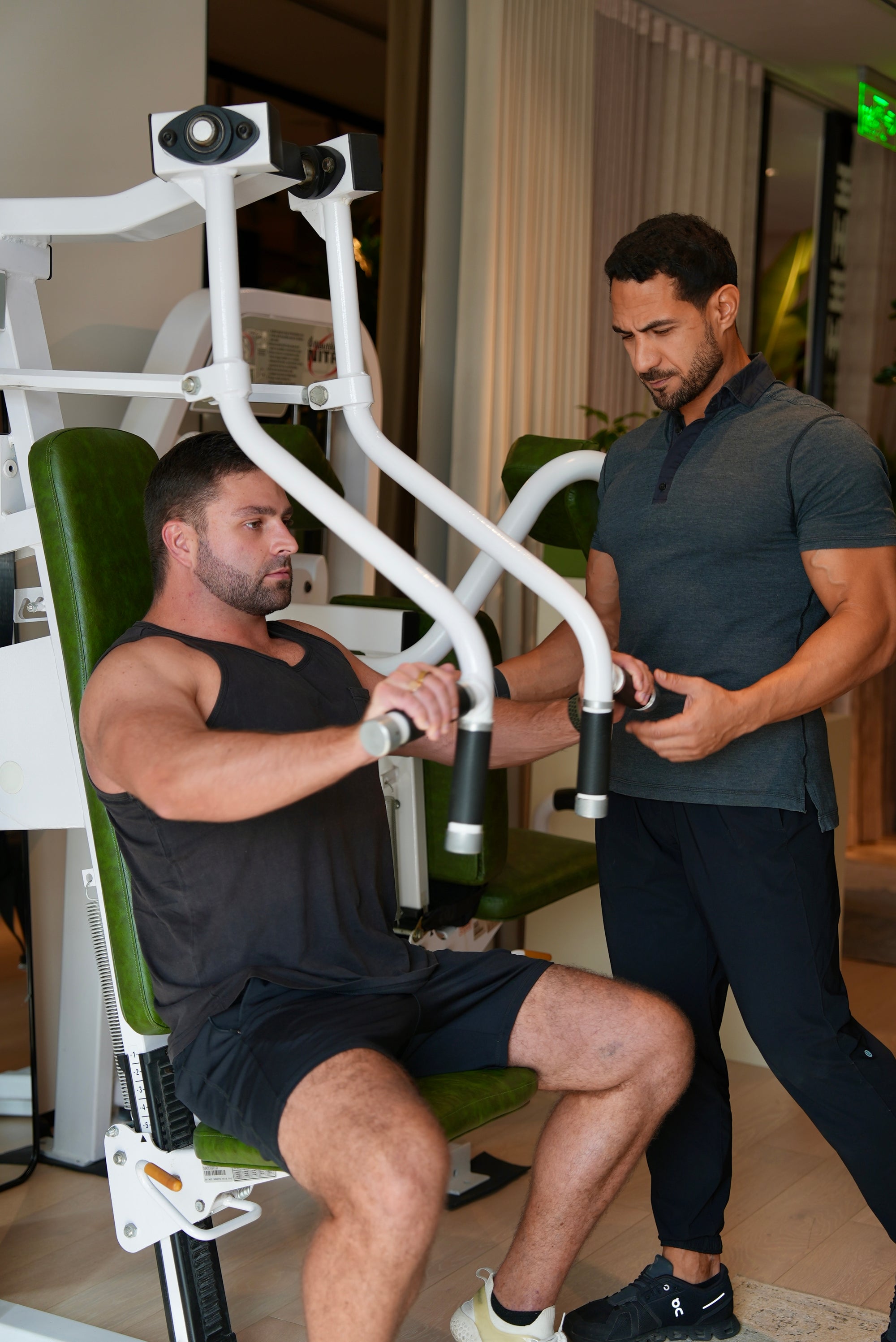 Man exercising on a weight machine with another man assisting him in a gym setting.