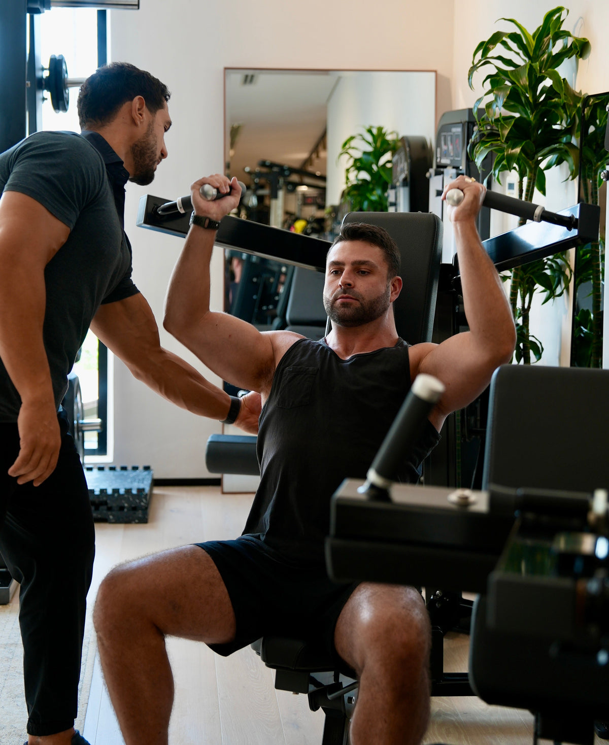 Personal trainer assisting a male client on a shoulder press machine during a strength training workout in Halm Collective Wellness Centre Dubai. Natural light, green plants, calm environment in a boutique gym.