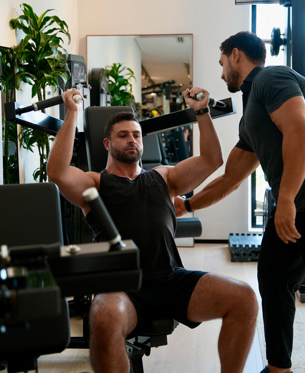 Personal trainer assisting a male client on a shoulder press machine during a strength training workout in Halm Collective Wellness Centre Dubai. Natural light, green plants, calm environment in a boutique gym.