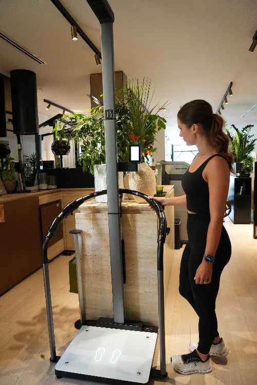 Female personal trainer standing next to a body composition machine adjusting the setting  with tropical plants and travertine in the background in private luxe boutique gym, Jumeriah Dubai. 